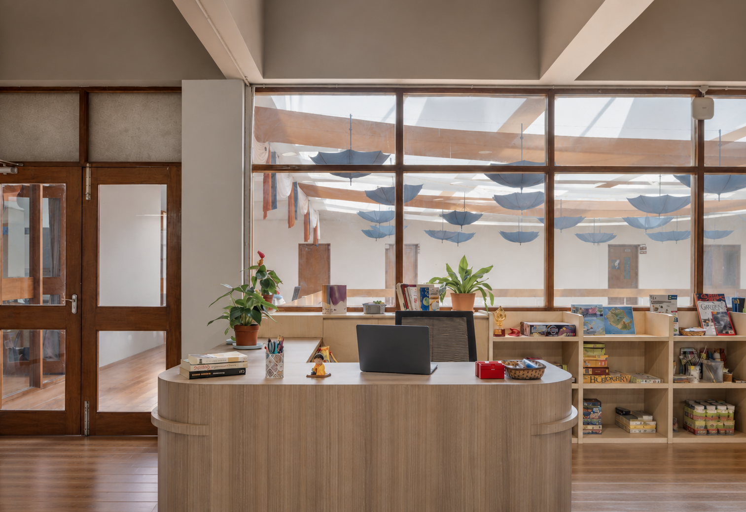 Reception desk in a light-filled institutional interior, with a curved timber counter, planting, bookshelves and large windows revealing suspended umbrella forms above